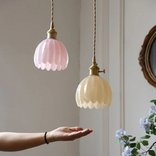 Two hanging pendant lights with scalloped glass shades, one pink and one beige, held by a hand against a neutral wall.